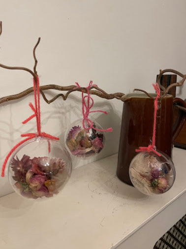 This image displays a set of pink dried flower baubles, elegantly suspended from a rustic branch. Each bauble is made from a clear, round globe, beautifully filled with an assortment of pink and purple dried flowers.
